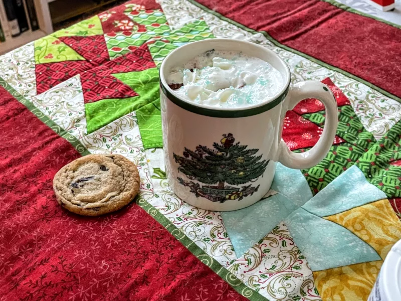 A mug of hot cocoa and a warm chocolate chip cookie sit on a holiday patterned table cloth