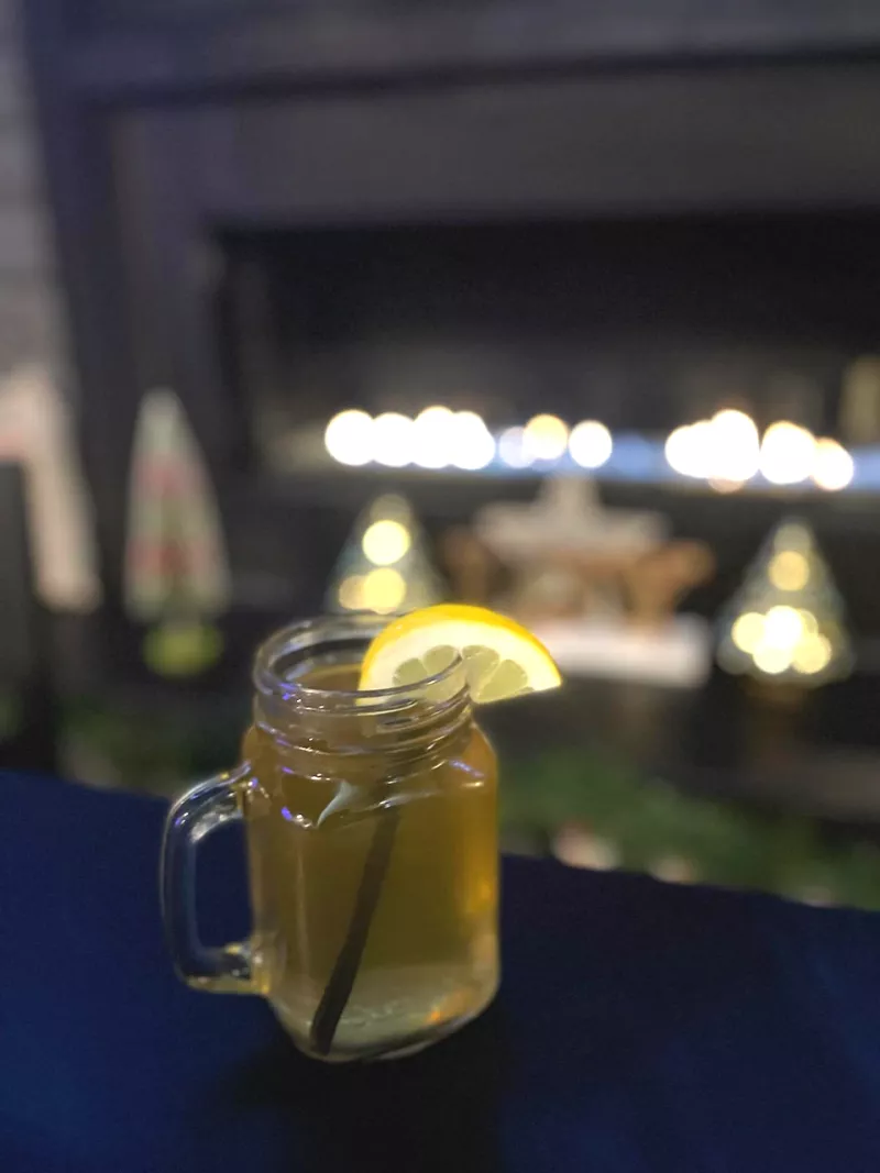 A mug of hot toddy sits on a table with a fire in the background.