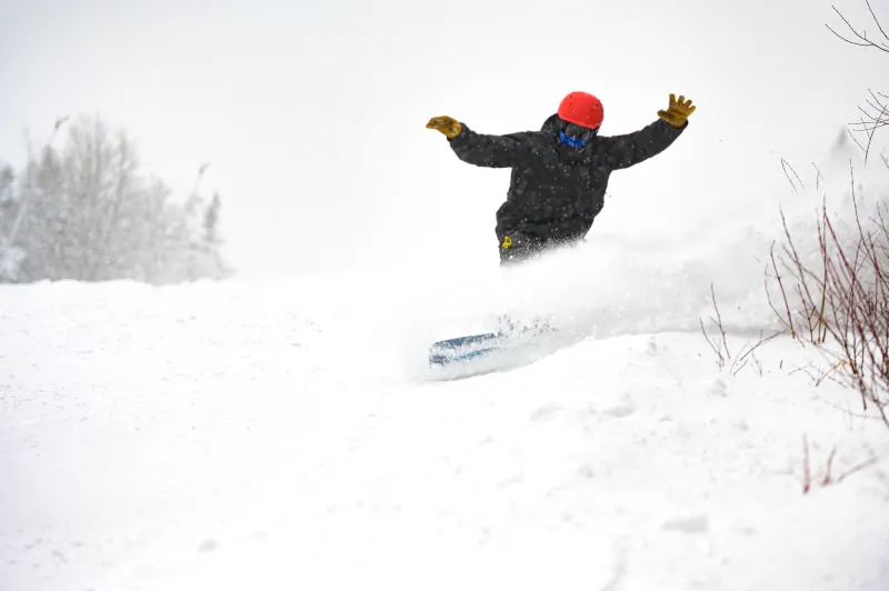 A snowboarder with a black jacket and red helmet ride through fresh powder on a snowy day.