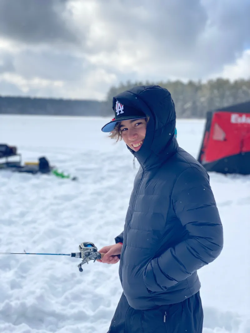 All bundled up, a boy smiles a the camera, her friends and fellow anglers in the background.