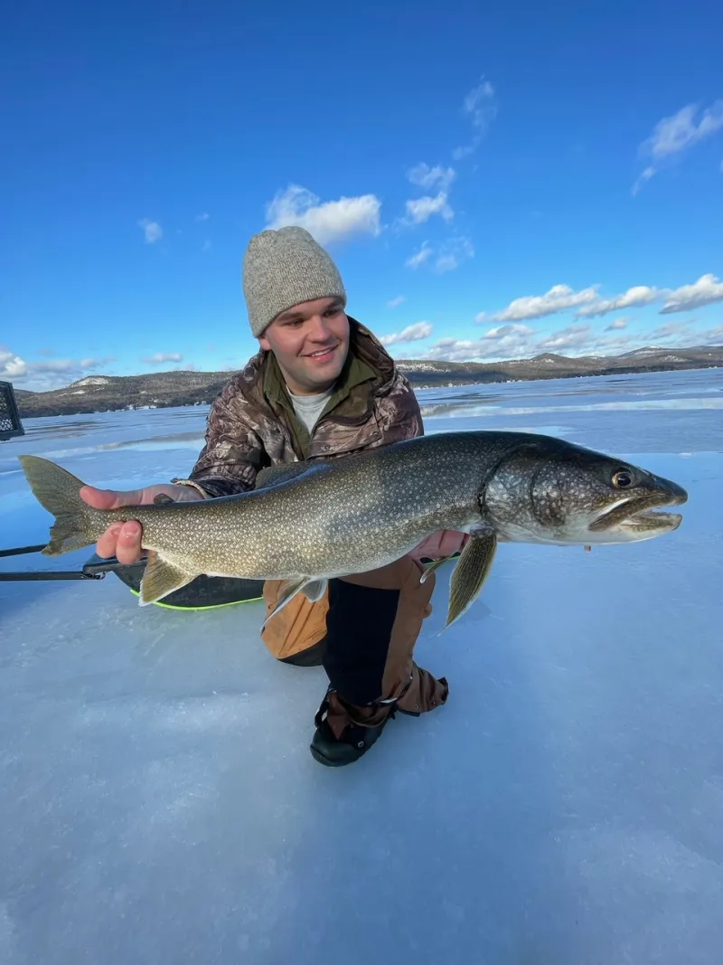 A man holds a massive lake trout out for the camera, kneeling on the ice.