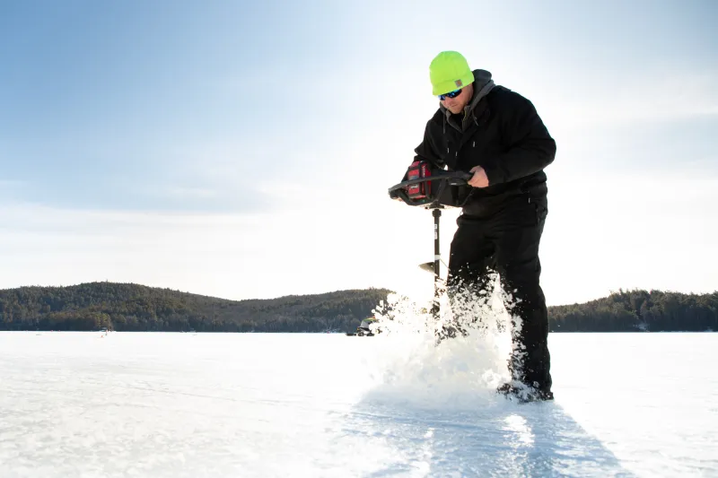 A man uses an electric auger to drill a hole in the ice on a sunny day.