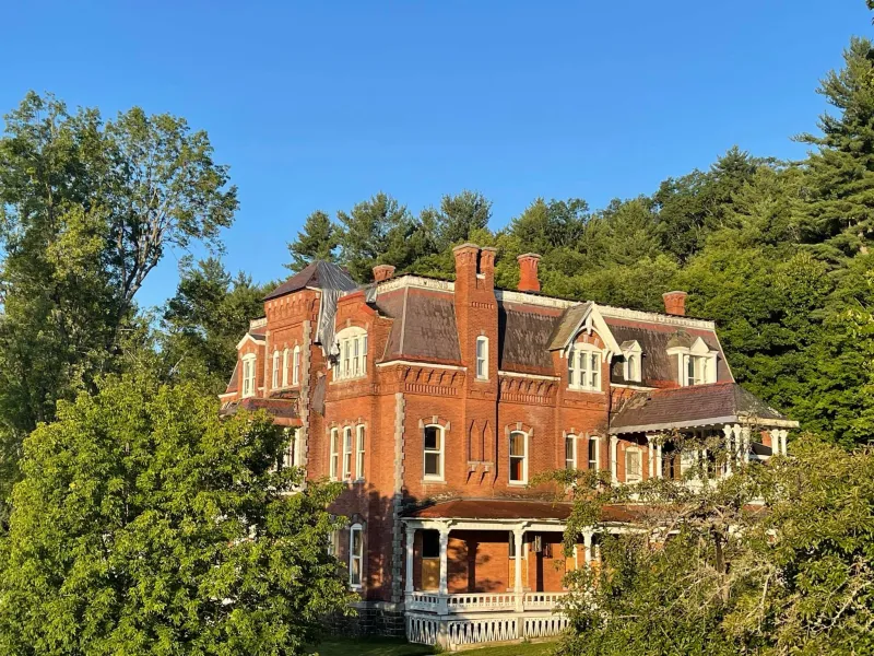 A three-story red brick mansion shines in the sun, surrounded by trees.