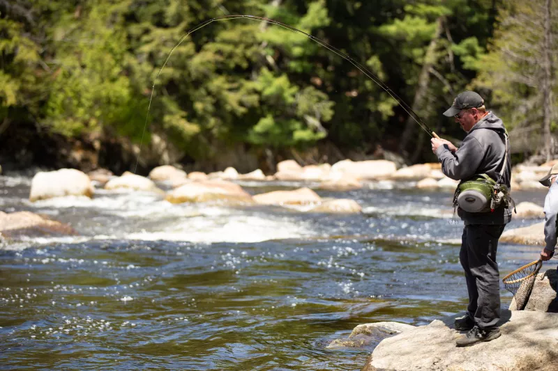 A fly fisherman reels his line in on the banks of a rocky river.