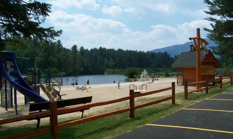A scenic shot of a calm lake with a playground and a wooden hut on the sandy beach and mountains in the background.