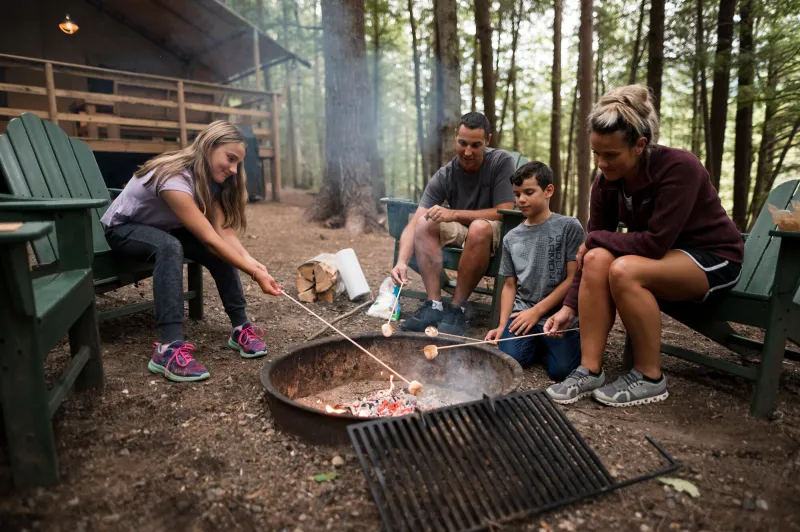 Two parents and their young kids roast marshmallows over a fire while sitting on Adirondack chairs in the woods.