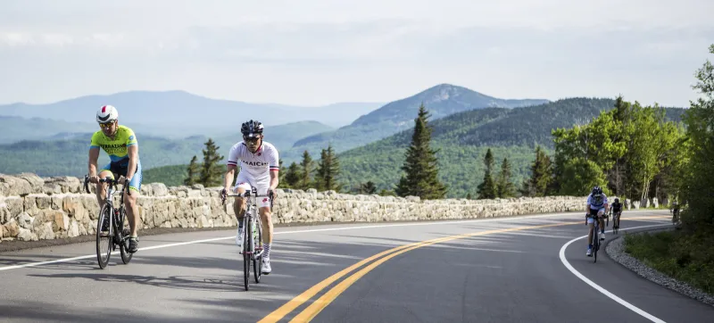 Two cyclists climb the toll road up Whiteface Mountain, with trees and mountains in the background.