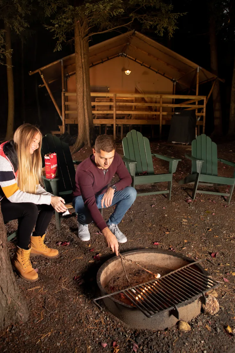 A couple sits at a campfire by Adirondack chairs and a glamping tent.