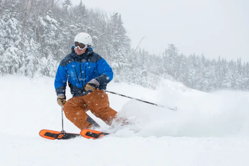 A skier in a blue coat and orange pants skis downhill during a snow storm.