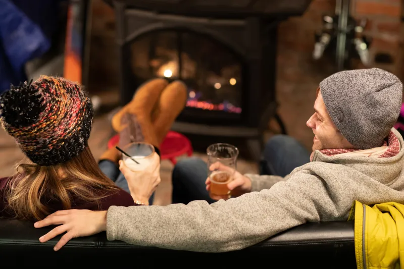 A couple sits by a fire holding cocktails and beer.