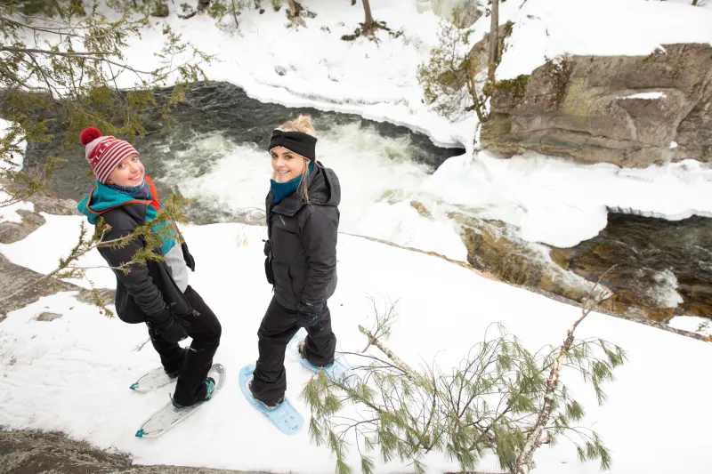 Two women look up at the camera from a snowy ledge above an icy river, while on snowshoes.