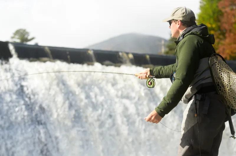 A person wearing a green jackets casts in front of a dam with a mountain in the backdrop