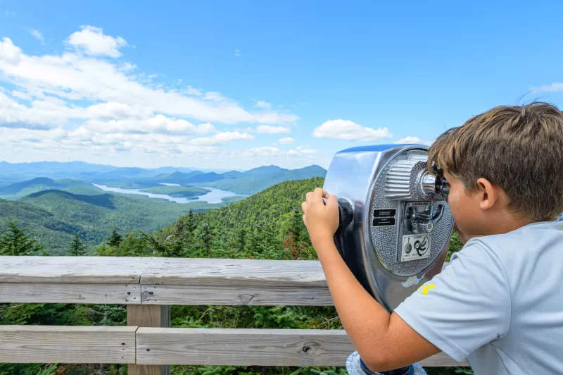 a boy looks out of the binoculars on an observation deck overlooking mountains.