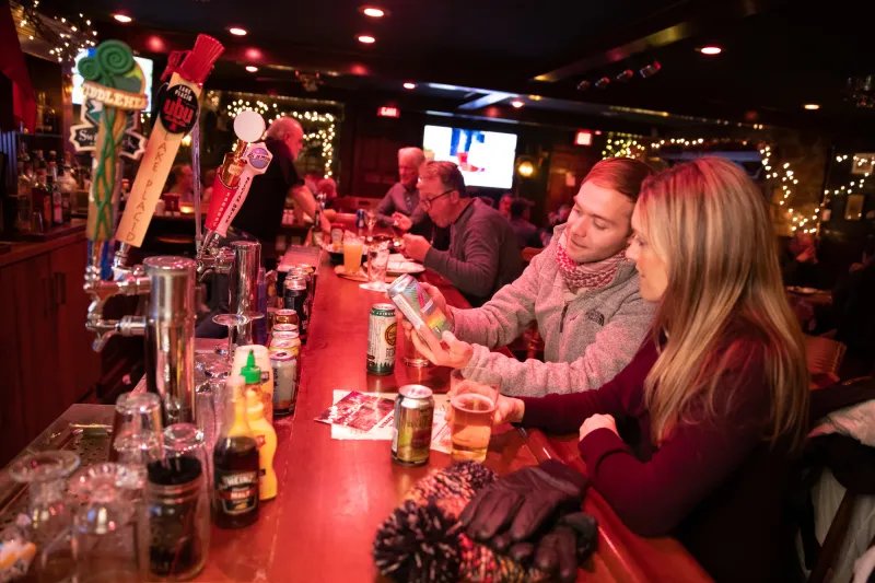 A man and woman look at a cellular phone together at the bar of a cozy, warmly lit pub.