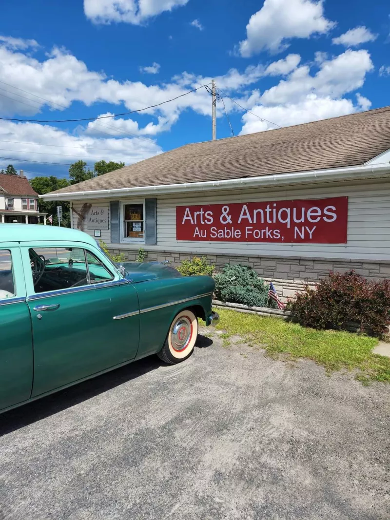 A bright teal vintage automobile sits parked outside a while building with a read sign that reads "Arts & Antiques."