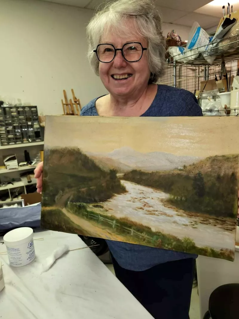 A cherubic woman with short white hair, glasses, and a big smile in her 50s holds an antique oil painting.