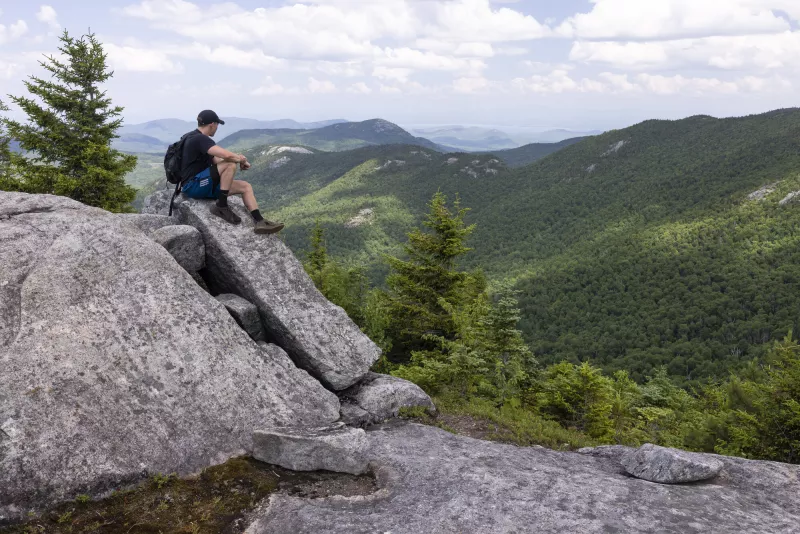 A hiker sits at elevation, on top of a glacial erratic, with an Adirondack mountain landscape in the distance