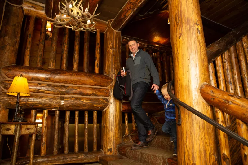 A man and toddler walk down grand log stairs in a lodge.