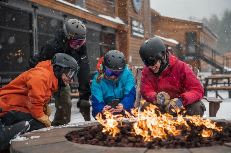 A group of skiers look at a phone around a fireplace.