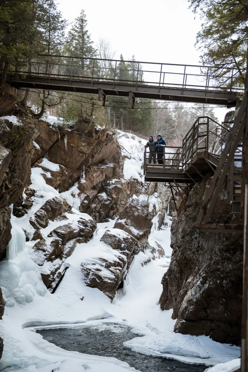 Two people stand on a hitch-up bridge along the stone cliff wall of High Falls Gorge and look down up on a waterfall during winter