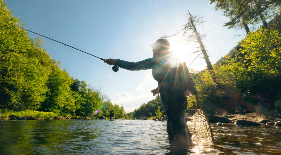 Angler stands in river casting a fly fishing line with sun shining behind them