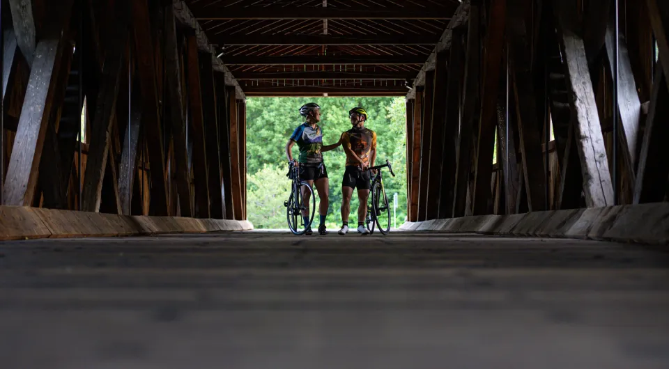 Two cyclists stand straddling their bikes inside of covered bridge