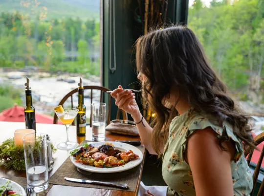 A woman with long dark hair eats a meal at a restaurant table with green trees and a river in the background.
