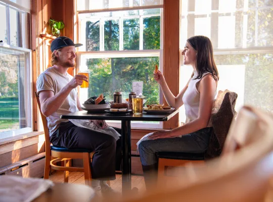 Male/female couple sits opposite each other at dining table in bright windowed room eating and drinking