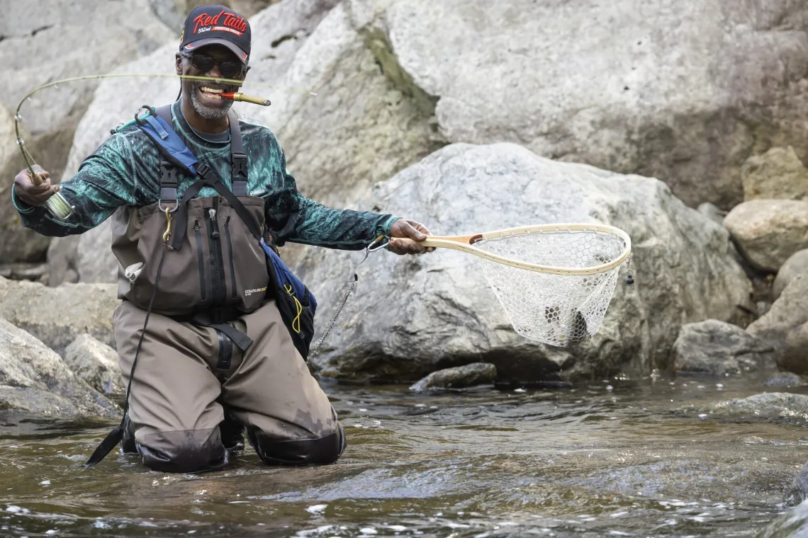 A grinning man with a cigar in his mouth holds a fly rod and net, showing off the trout he caught.