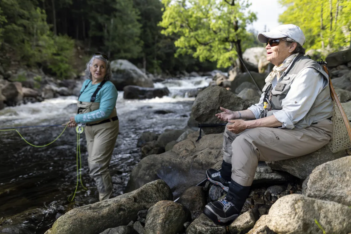 Two women in waders enjoying Adirondack fly fishing. One stands in the river fishing, the other perches on a riverside rock.