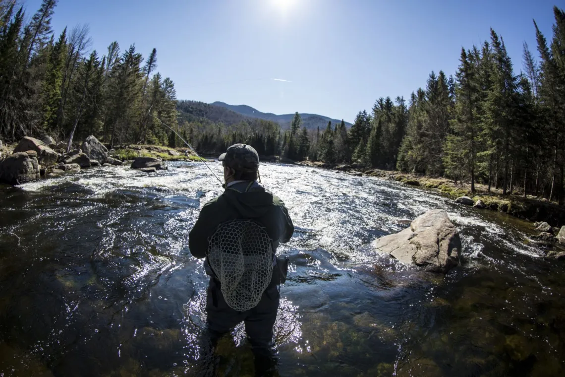 A man wading in the Ausable river fly fishes with mountain peaks in the background