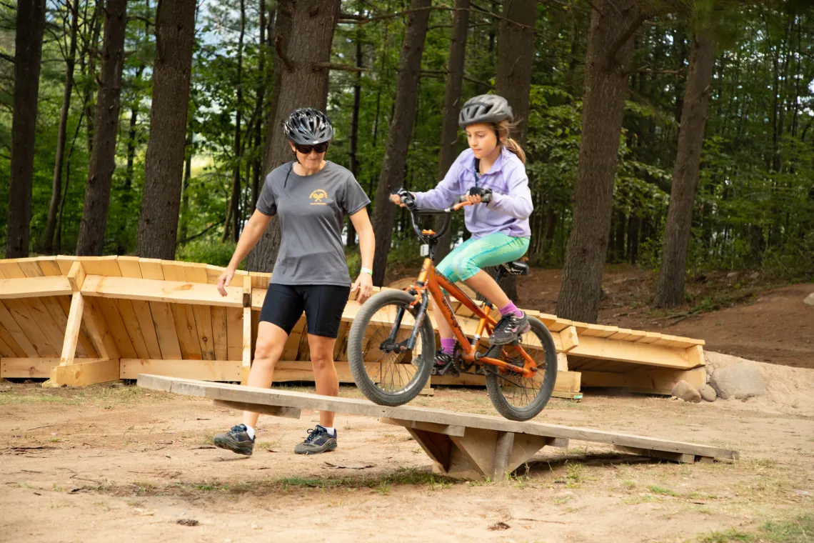 A mother helps her daughter on a wooden bike park feature